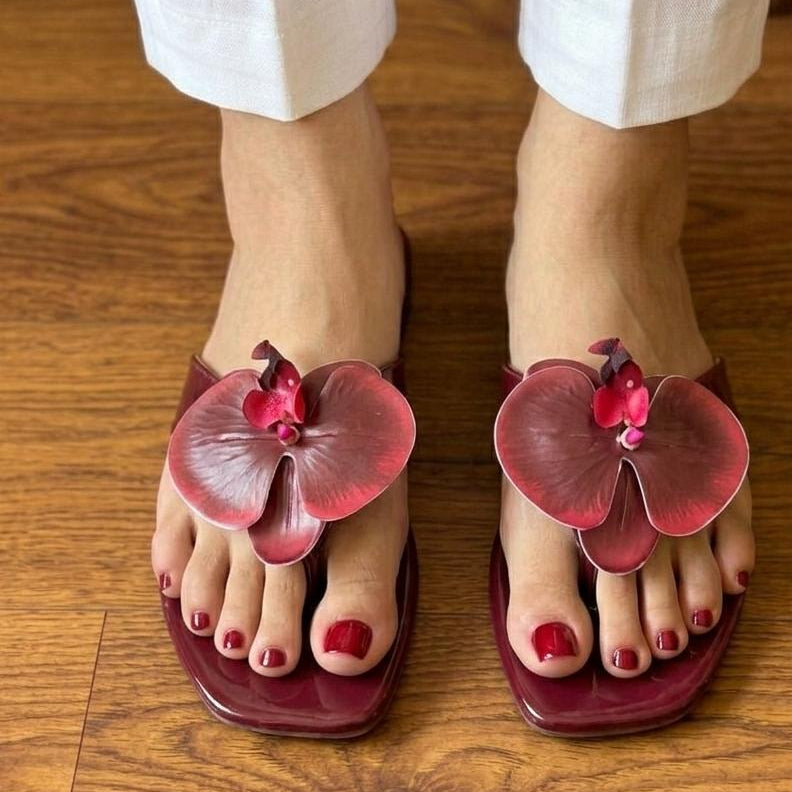 Red floral sandals worn on feet with white pants on a wooden floor.