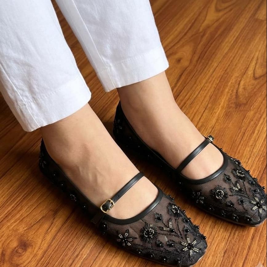 Black embellished shoes worn with white pants on a wooden floor