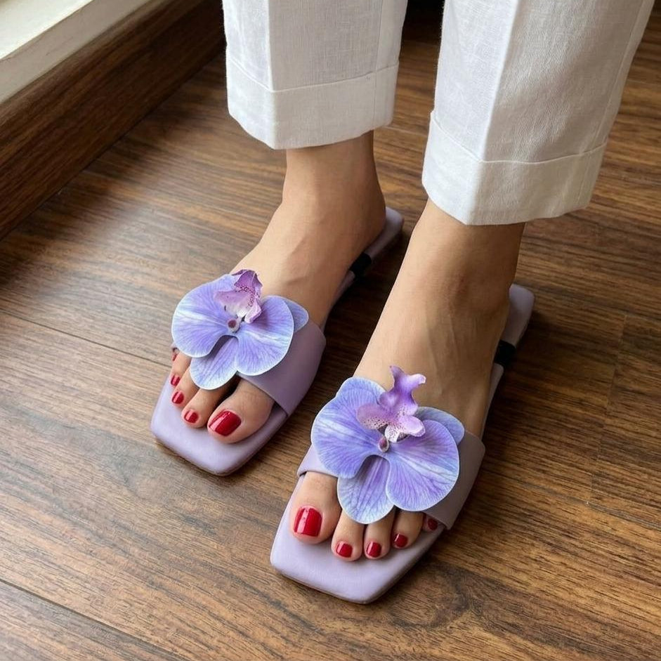 Purple sandals with floral design worn indoors on a wooden floor.