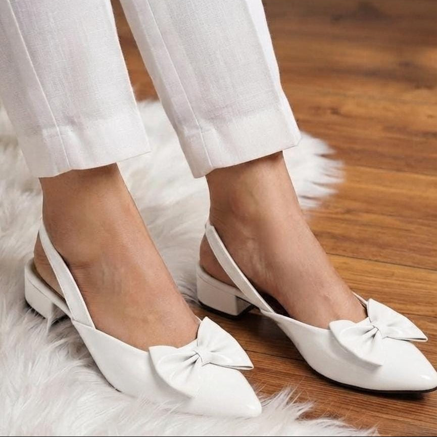 White slingback pointed-toe flats with large bow details, shown on a model wearing white cropped pants on a wooden floor with a white faux-fur rug.