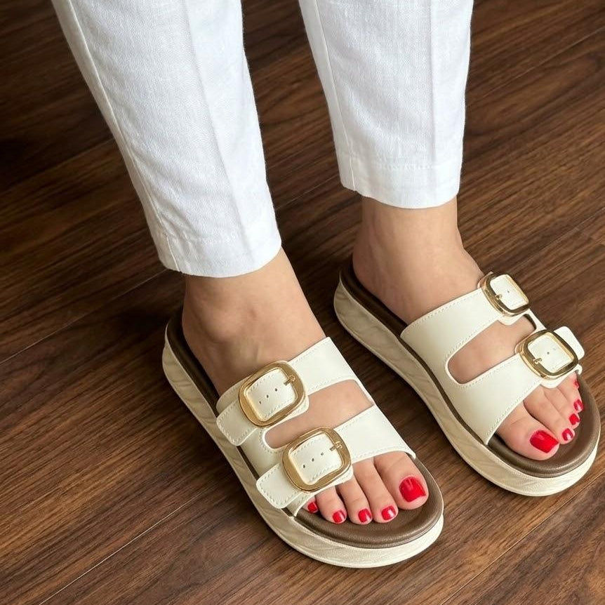 White sandals with gold buckles worn on a wooden floor
