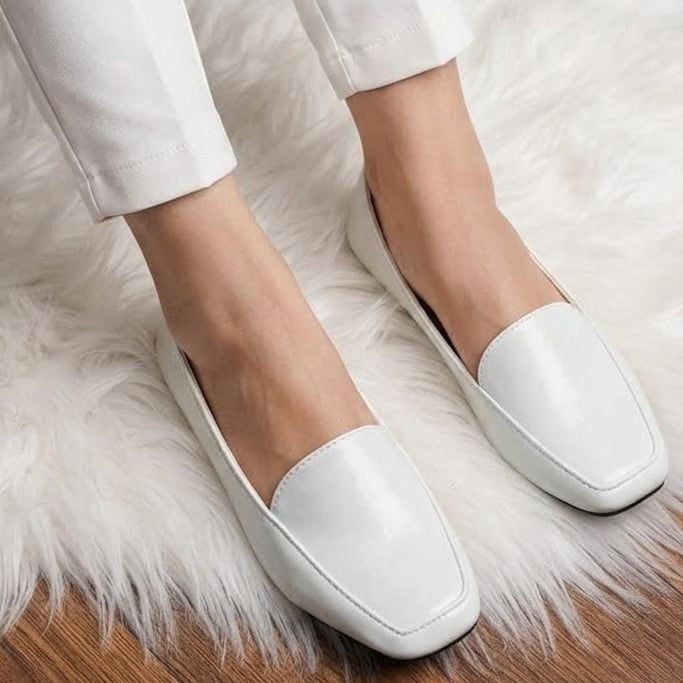 White leather loafers with a square toe, worn with white cropped trousers on a white faux fur rug over a wooden floor.