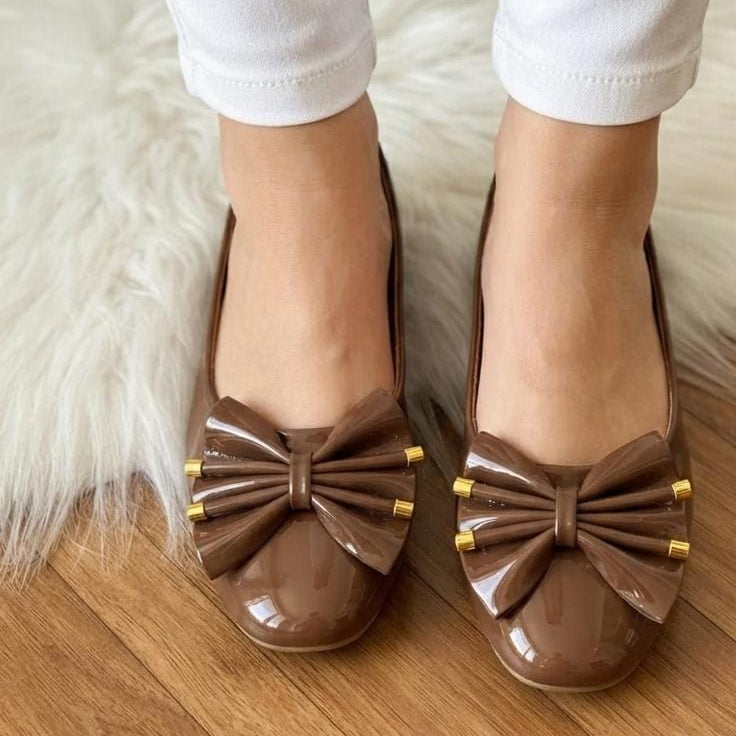 A pair of brown patent leather ballet flats with a large bow and gold-tone hardware, shown on a model wearing white pants on a wooden floor with a white faux fur rug.