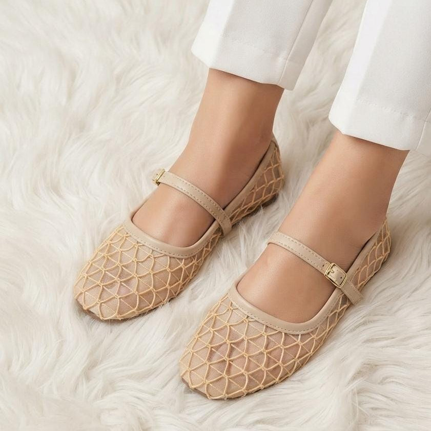 Beige open-knit Mary Jane flats with ankle straps and small gold buckle details, shown on a model's feet against a white faux fur rug.