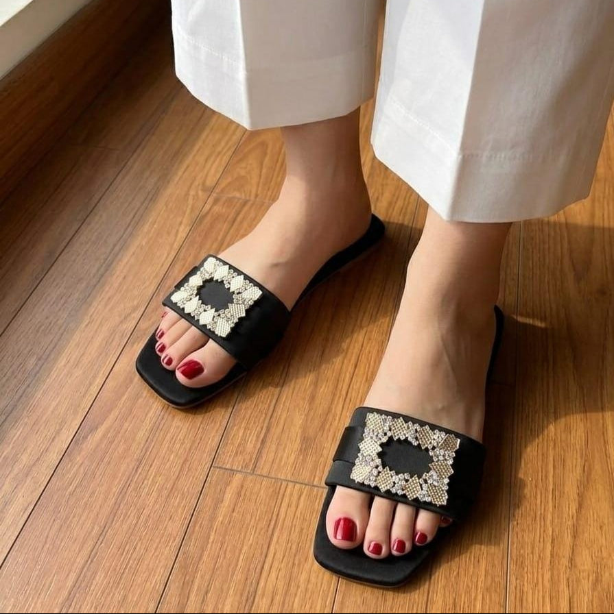 A person wearing black slide sandals with ornate square embellishments on the straps, shown on a wooden floor with white cropped pants visible.