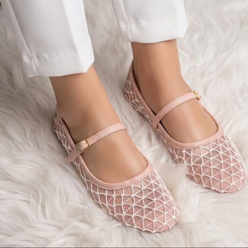 A close-up of a person wearing light pink openwork mesh Mary Jane flats with a small gold buckle, paired with white pants, standing on a white faux fur rug.
