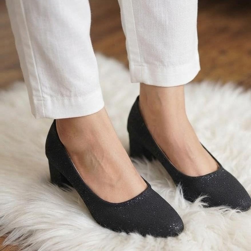 A person wearing black pointed-toe low-heel pumps with white pants, standing on a white faux-fur rug over a wooden floor.