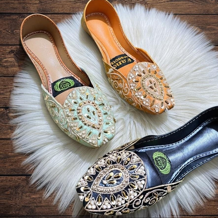 Three embellished flat mule shoes arranged on a white faux-fur rug over a dark wood surface. The shoes have ornate embroidered and jeweled details: one in pale mint green, one in tan, and one in black.