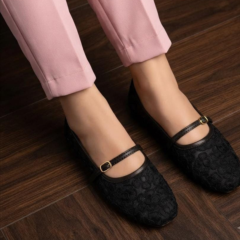 A person wearing black textured Mary Jane flats with gold-tone buckle straps, paired with light pink trousers, standing on a dark wood floor.