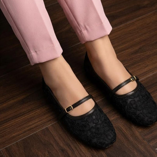 A person wearing black textured Mary Jane flats with gold-tone buckle straps, paired with light pink trousers, standing on a dark wood floor.