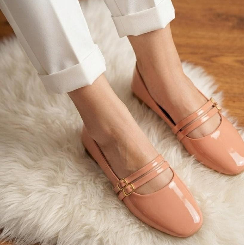 A person wearing light peach patent leather Mary Jane flats with gold buckle details, paired with white cuffed trousers, standing on a white faux fur rug over a wooden floor.