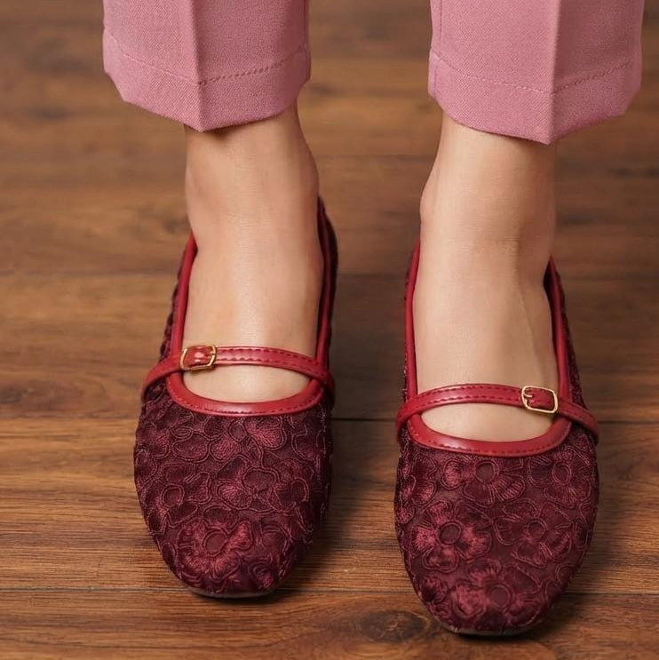 A close-up of a person wearing burgundy lace flat shoes with red straps and gold buckle details, paired with pink pants, standing on a wooden floor.