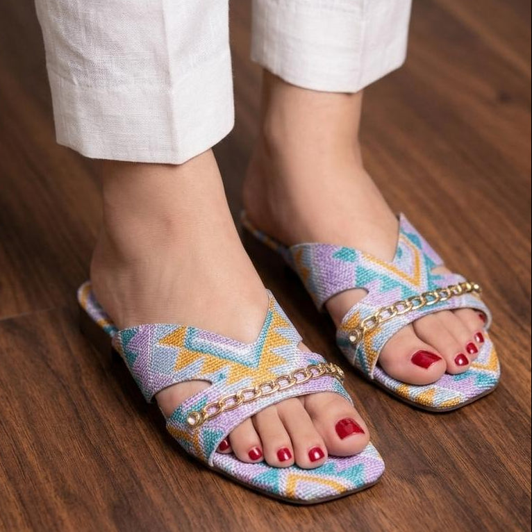 Colorful patterned sandals worn with white pants on a wooden floor.
