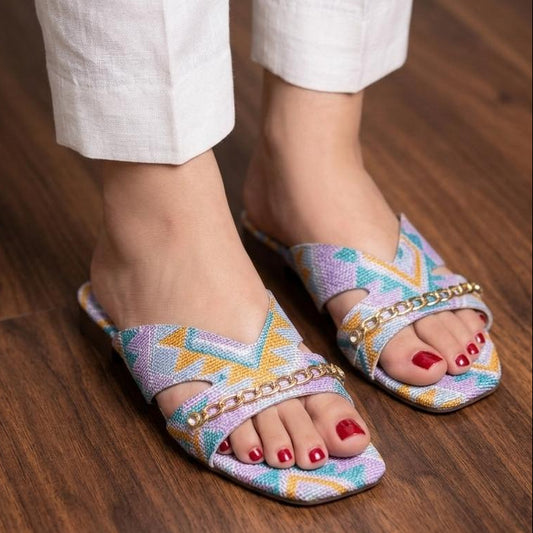 Colorful patterned sandals worn with white pants on a wooden floor.