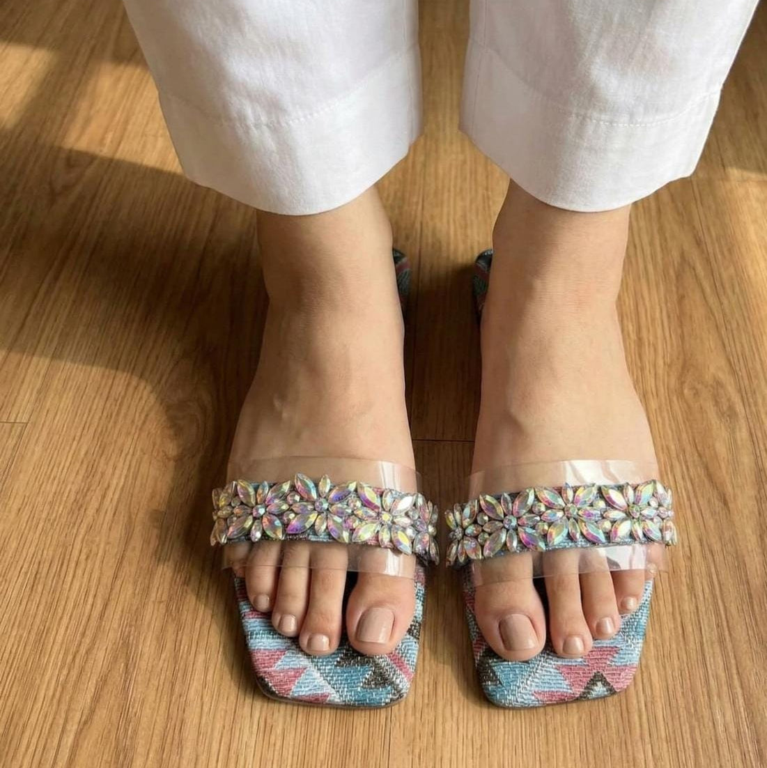 A close-up of a person's feet wearing clear slide sandals with iridescent floral embellishments across the toes and patterned blue, pink, and green square toes, standing on a wooden floor.