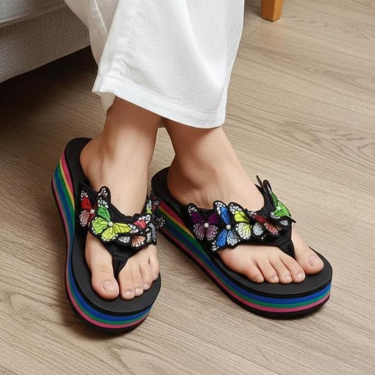 Black sandals with colorful butterfly decorations worn indoors on a wooden floor.