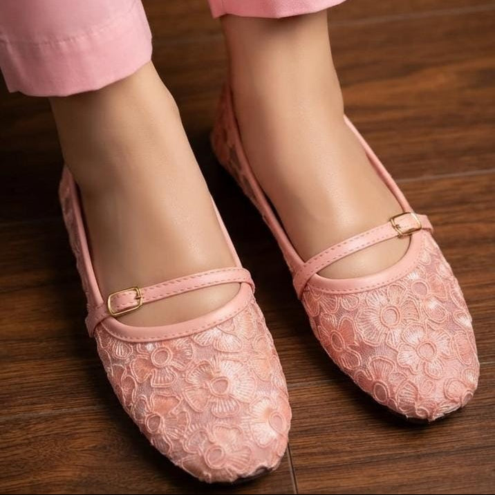 A close-up of a person wearing pink lace ballet flats with a small gold buckle, paired with pink pants, standing on a dark wood floor.