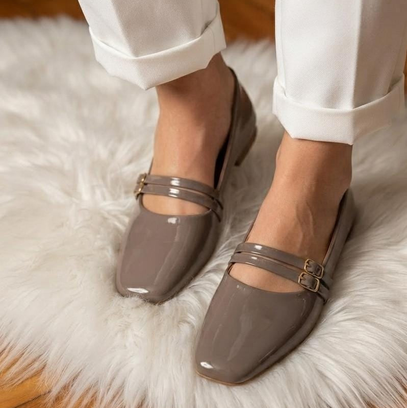 A person wearing glossy taupe Mary Jane flats with double straps and gold-tone buckles, standing on a white faux-fur rug over a wooden floor.