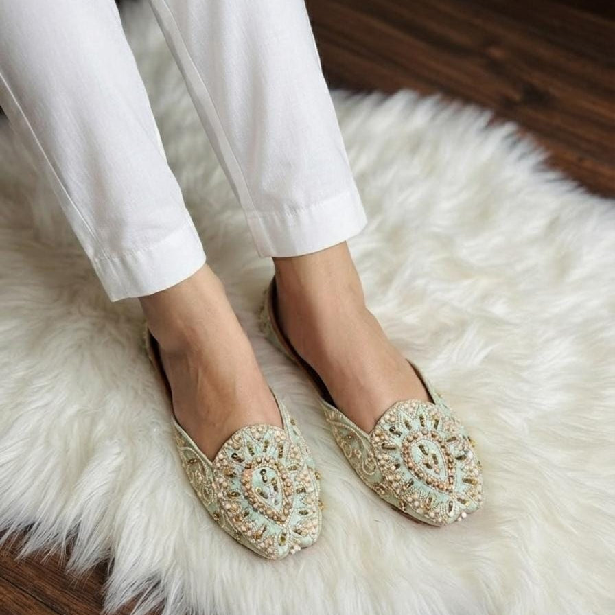 A person wearing white pants and light beige embellished flat shoes with intricate beadwork, standing on a white faux fur rug over a dark wood floor.