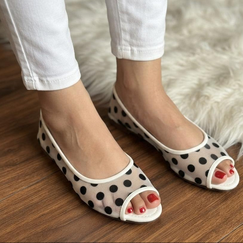 A person wearing white pants and beige slip-on peep-toe flats with black polka dots, standing on a wooden floor next to a white faux fur rug.