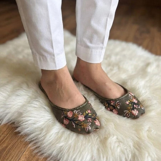 A person wearing white pants and olive green pointed-toe flats with a pink floral pattern, standing on a white faux fur rug over a wooden floor.