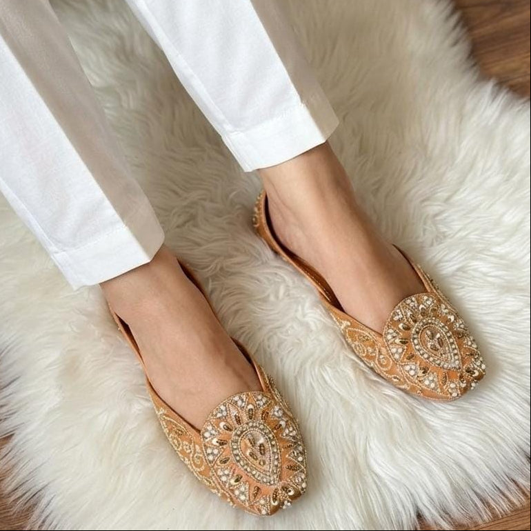 A person wearing white trousers and ornate beige flat shoes with intricate gold and silver beadwork, sitting on a white faux fur rug over a wooden floor.