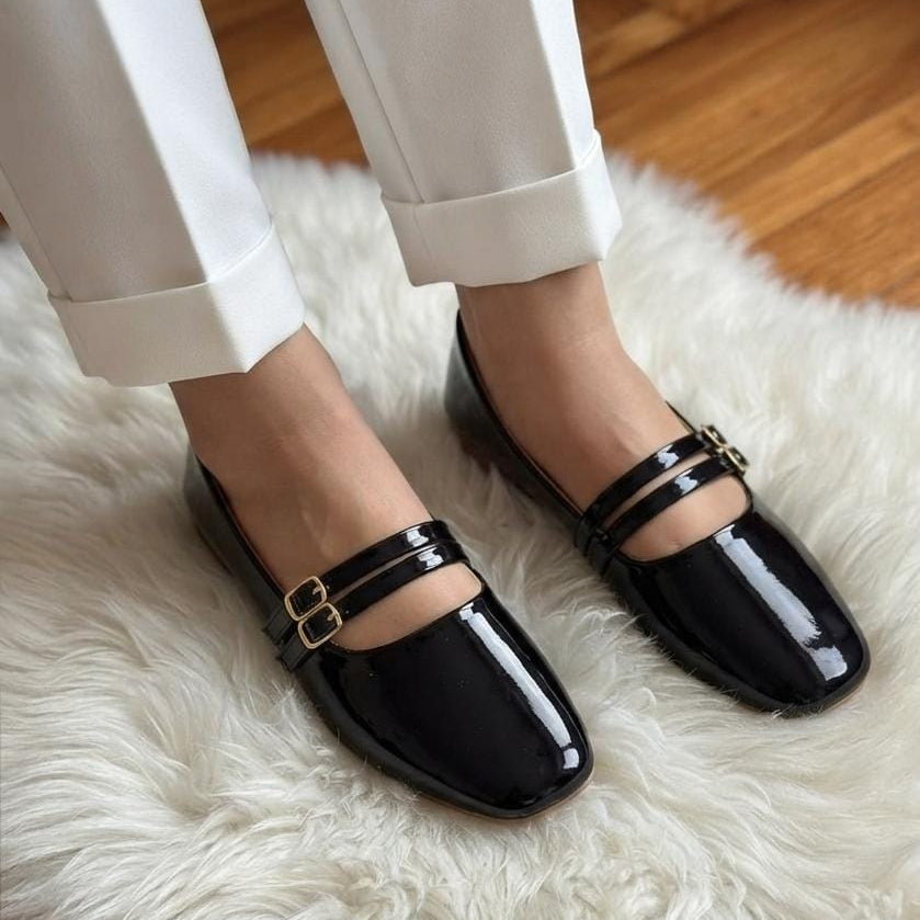 A person wearing glossy black patent leather Mary Jane flats with double straps and gold-tone buckles, paired with white pants, sitting on a white faux fur rug over a wooden floor.