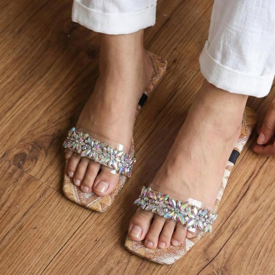 A close-up of a person wearing clear rhinestone-embellished slide sandals with a beige woven-texture sole, shown on a wooden floor with white cuffed pants visible.