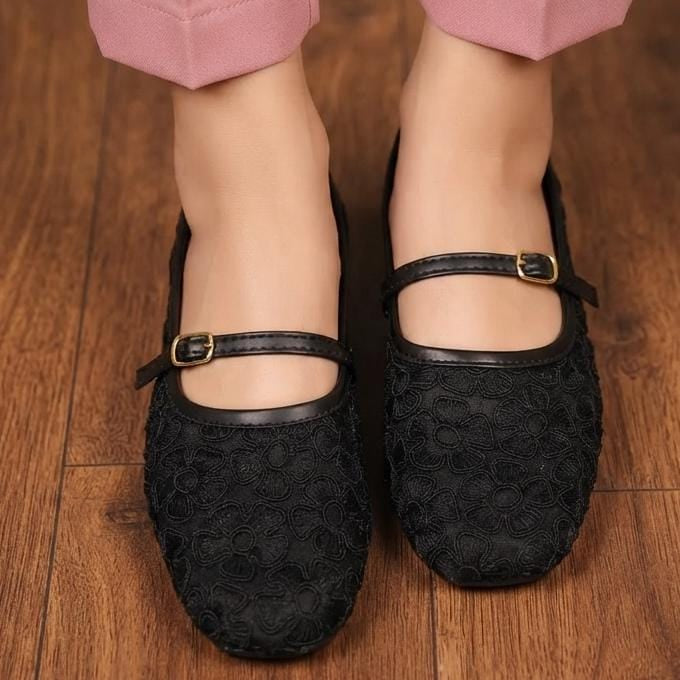 A close-up of a person wearing black textured Mary Jane flats with gold buckle straps, paired with pink pants, standing on a wooden floor.