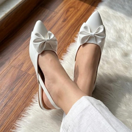 White pointed-toe shoes with bows worn by a person on a wooden floor.