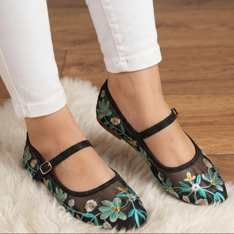 A person wearing black Mary Jane flats with teal, white, and yellow floral embroidery on a white faux fur rug over a wooden floor, paired with white pants.