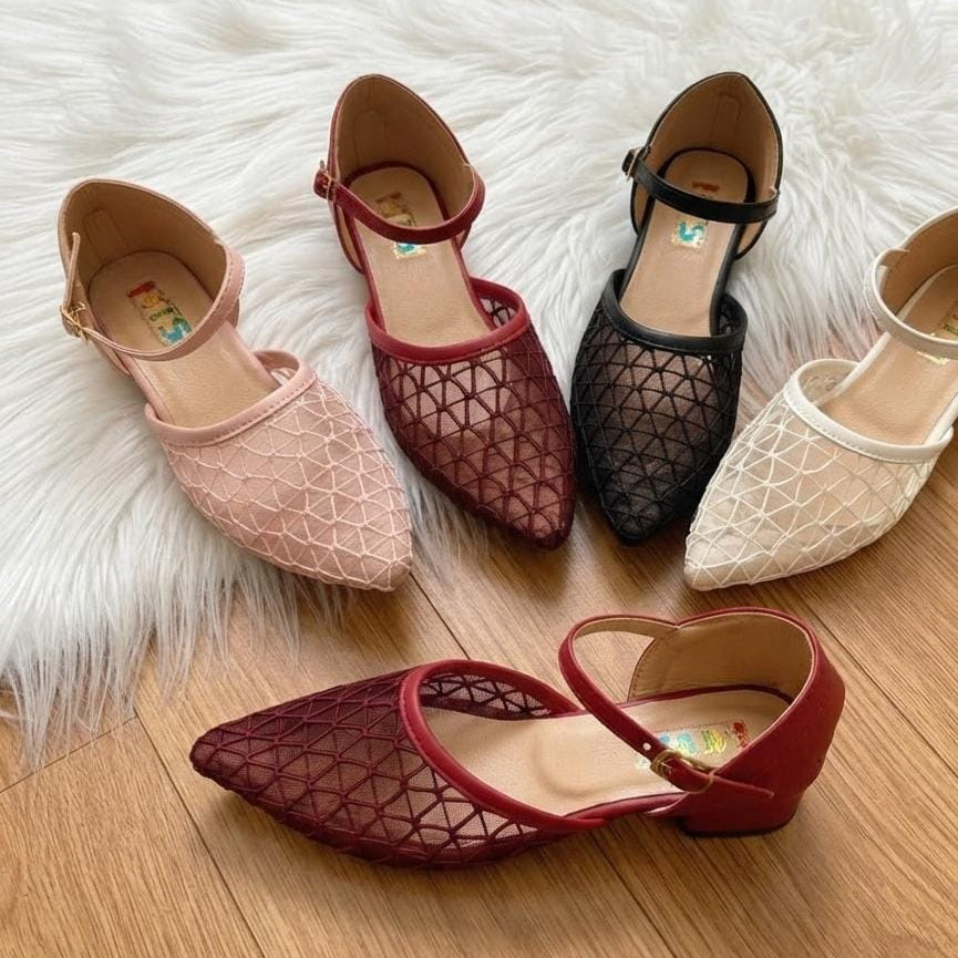 Four women's low-heeled flats arranged on a white faux-fur rug over a wood floor. The shoes have a geometric openwork upper and ankle straps, shown in blush pink, burgundy, black, and off-white.