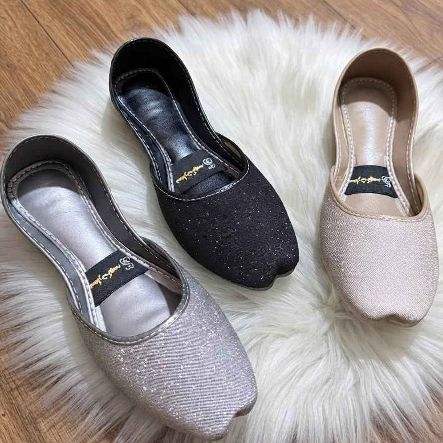 Three glittery slip-on flats in silver, black, and nude beige, arranged on a white faux fur rug over a wooden floor.