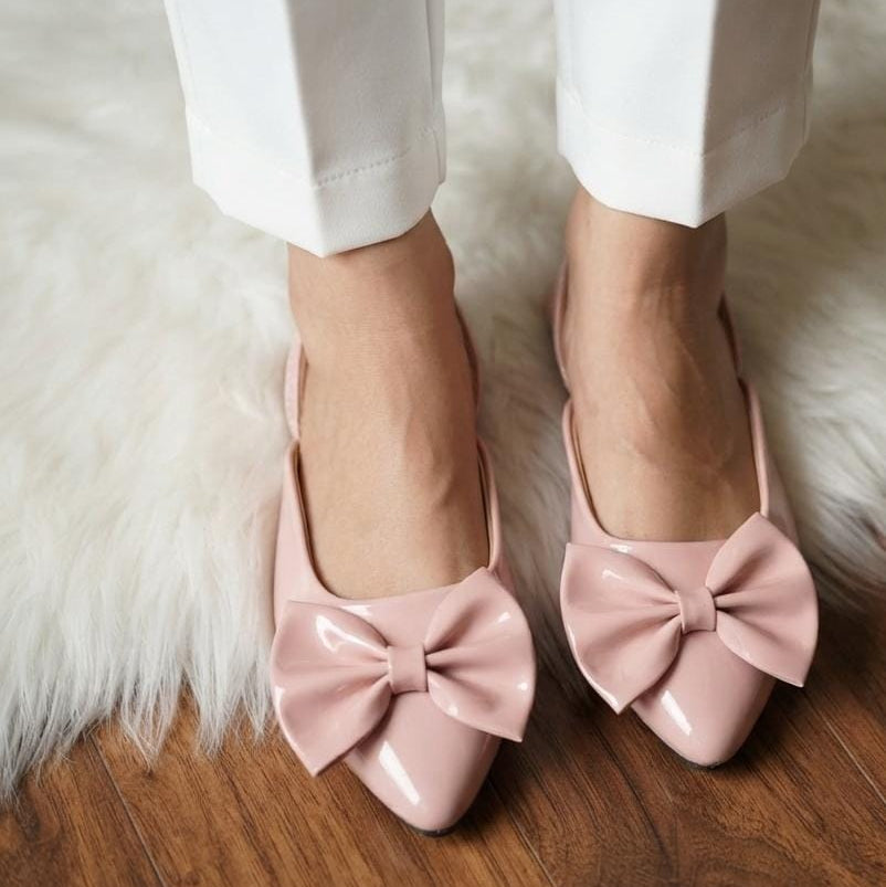 A close-up of a person wearing glossy light pink pointed-toe flats with large bow details, paired with white pants, standing on a wooden floor over a white faux-fur rug.