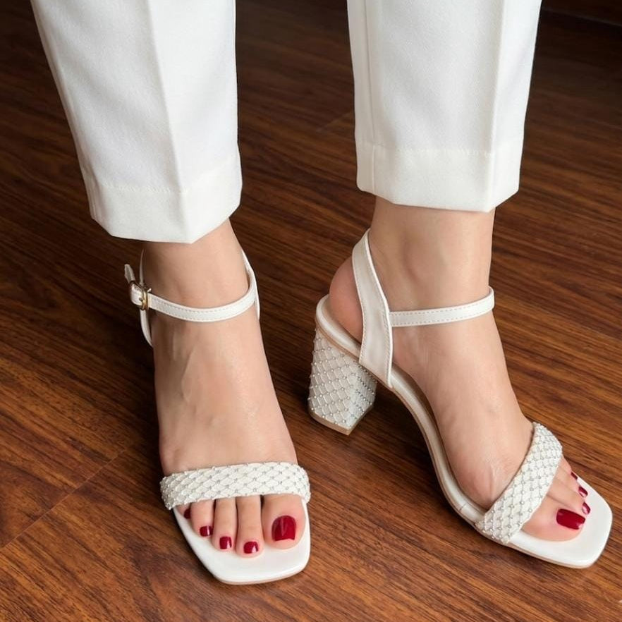 White woven-look block-heel sandals with ankle straps and square toes, shown on a model wearing white pants on a wooden floor.