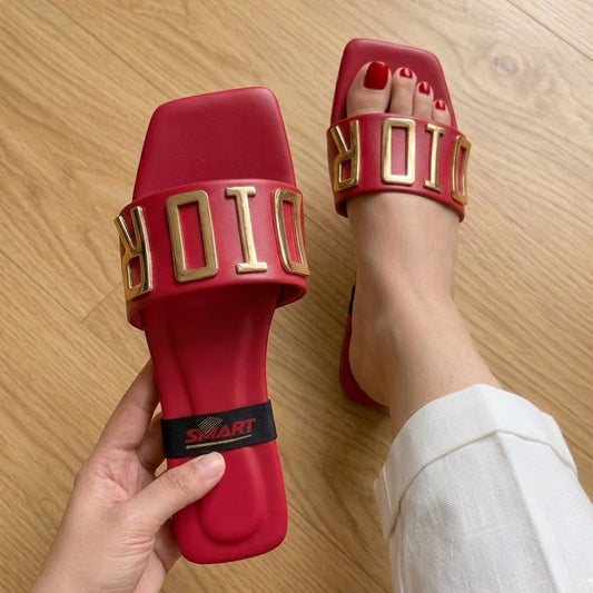 Red slide sandals with gold letter 'ORIO' strap details, shown with one being held by a hand and the other worn on a foot with red toenail polish, on a light wood floor.