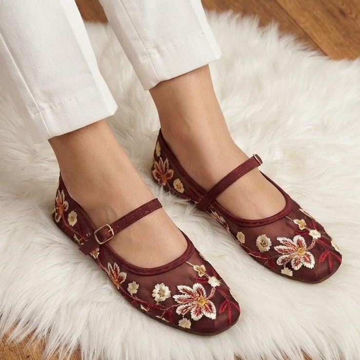 A person wearing burgundy flat shoes with floral embroidery and Mary Jane straps, standing on a white faux fur rug over a wooden floor.