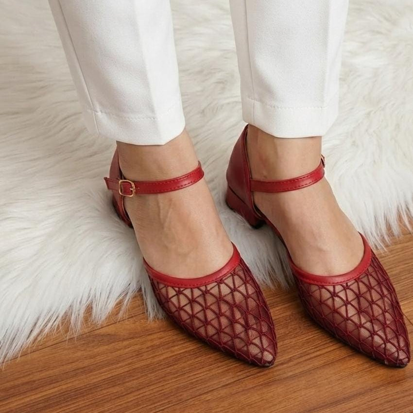A person wearing red pointed-toe Mary Jane flats with a geometric openwork upper, paired with white pants, standing on a wooden floor over a white faux-fur rug.