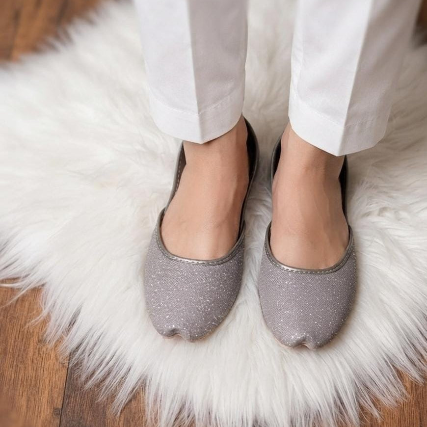 A person wearing white pants and silver glitter flat shoes standing on a white faux fur rug over a wooden floor.