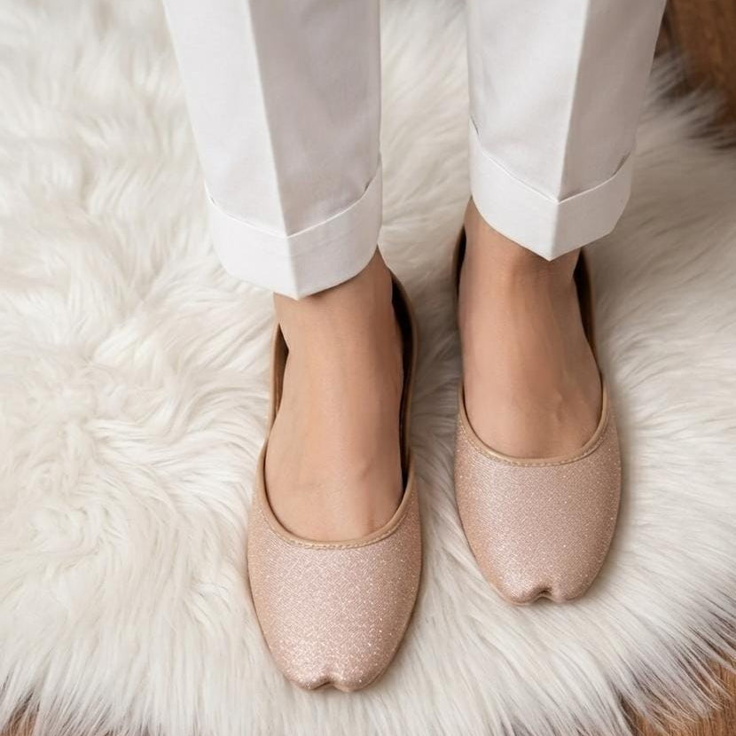 A close-up of a person wearing white pants and sparkling nude-pink ballet flats, standing on a white faux fur rug.