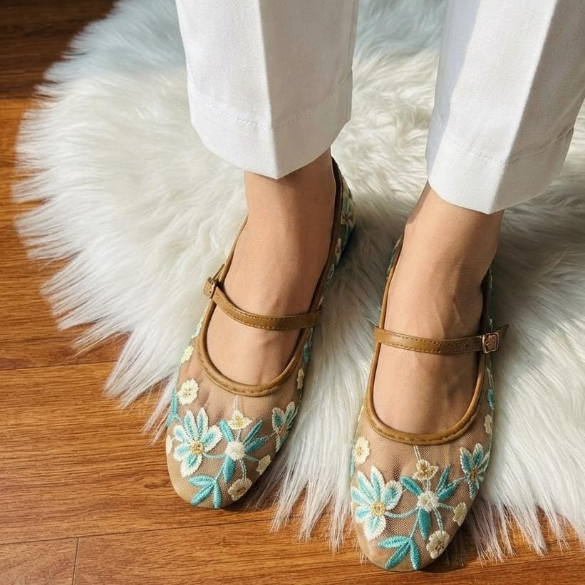 A person wearing white pants and tan Mary Jane flats with turquoise and cream floral embroidery, standing on a white faux-fur rug over a wooden floor.