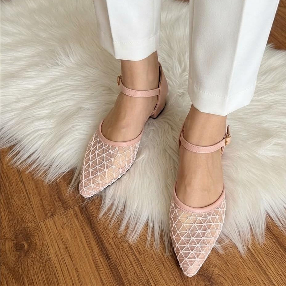 A person wearing light pink pointed-toe flats with a geometric mesh pattern and ankle straps, standing on a white faux-fur rug over a wooden floor.
