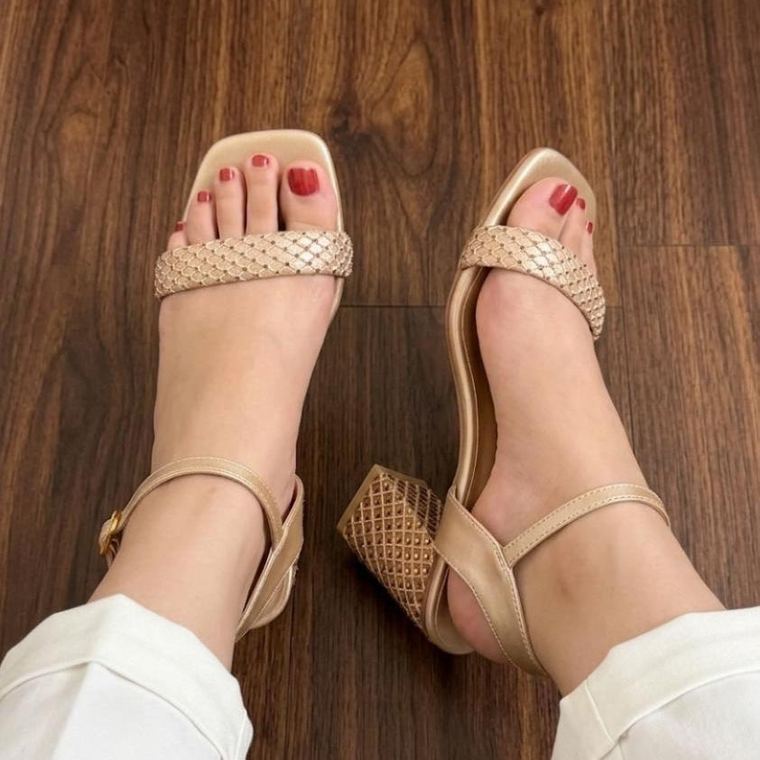 A pair of women's block-heel sandals in a light beige/tan color with woven textured straps, shown being worn on feet with white pants on a dark wood floor.