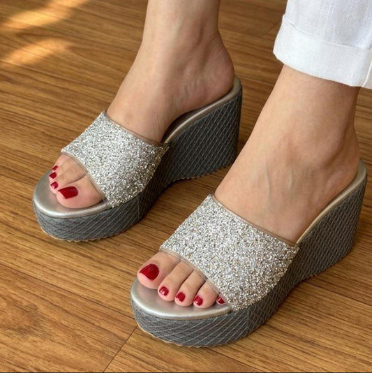 A close-up of a person wearing silver glitter platform mule sandals with a textured gray wedge heel, paired with white cropped pants, standing on a wooden floor.