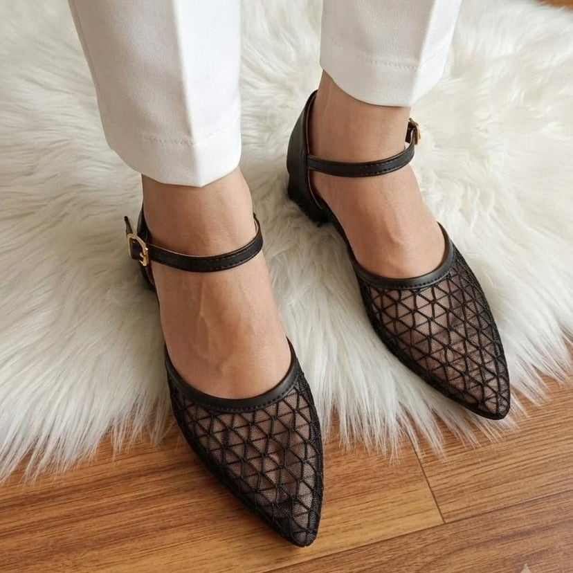 Black pointed-toe Mary Jane flats with a geometric mesh pattern, worn with white pants on a white faux fur rug over a wooden floor.