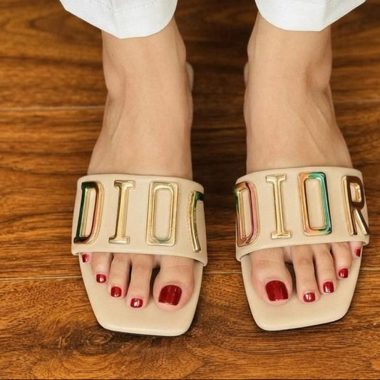 A close-up of a person wearing beige Dior slide sandals with metallic gold, green, and brown lettering on the straps, standing on a wooden floor.