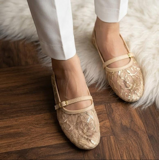 A close-up of a person wearing beige floral-embroidered ballet flats with a thin strap and small gold buckle, paired with white trousers, standing on a dark wood floor over a white faux-fur rug.