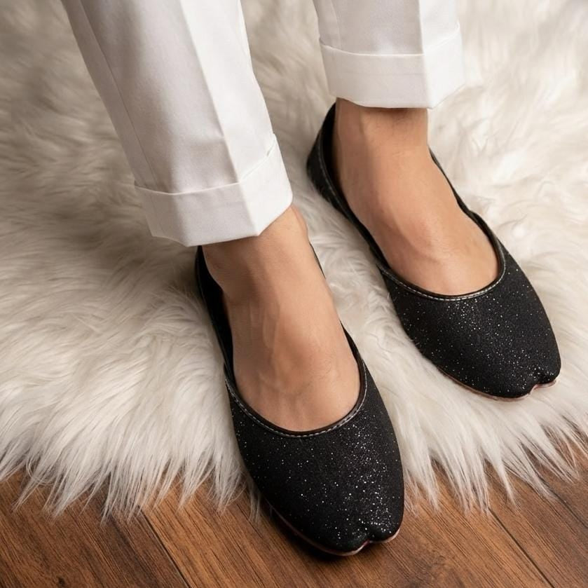Black glitter flat shoes with a traditional tabi-style toe, worn with white trousers on a white faux fur rug over a wooden floor.