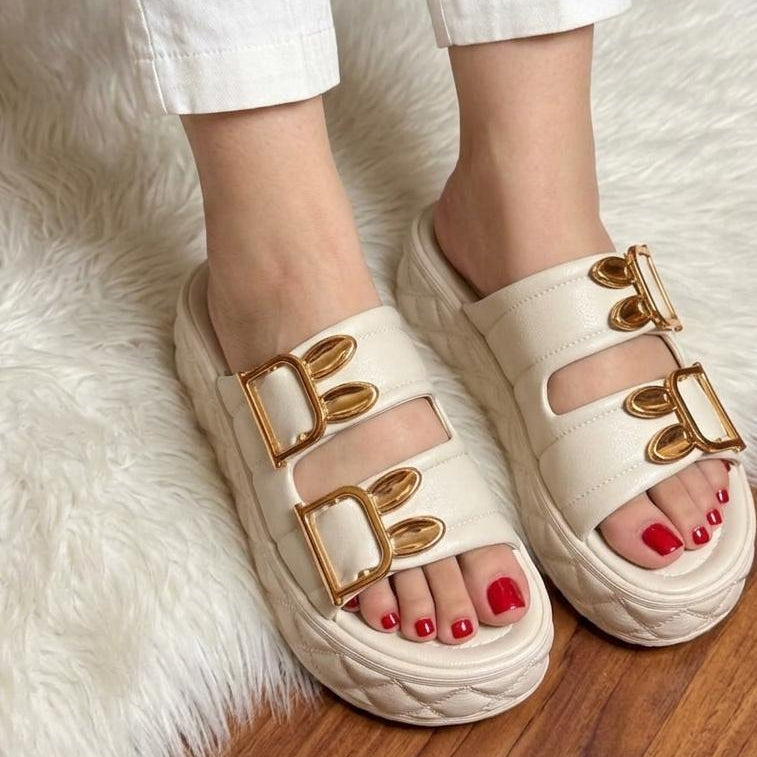 White sandals with gold buckles worn on a fluffy white rug.