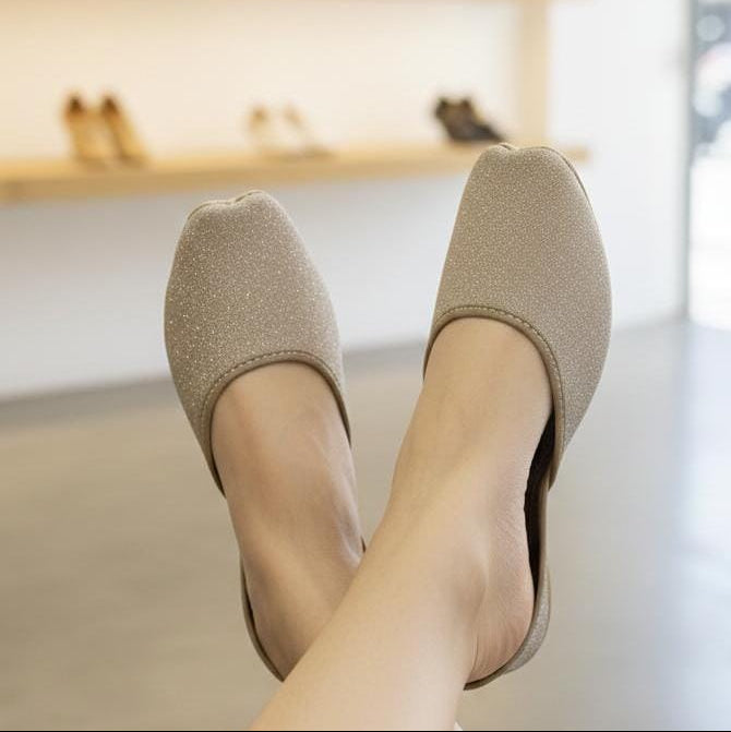A person wearing a pair of beige glitter slip-on mule shoes, with their legs crossed. The background shows a softly blurred shoe store interior with wooden shelves.